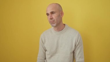 Gülümseyen, kendine güveni tam kel bir adam, kolları çapraz ve elleri çenesinde, bir süveter içinde, biraz mutluluk dolu, sarı izole bir arka planda gururla duruyor. Olumlu düşüncenin bir resmi.