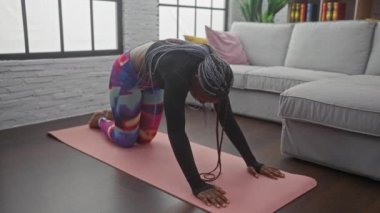 Aktif giyinmiş Afrikalı bir kadın modern bir oturma odasındaki paspasın üzerinde yoga yapıyor..