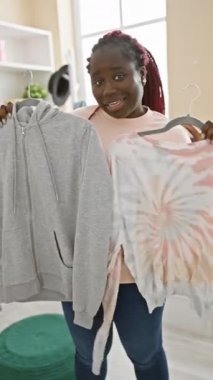 Saçları örgülü gülümseyen genç bir kadın, şık giyinme odasında kendine güveni ve tarzı olan kıyafetler seçer..