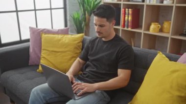 Modern bir oturma odasında renkli yastıklar ve kitaplıklarla dizüstü bilgisayarla çalışan sakallı genç bir İspanyol..