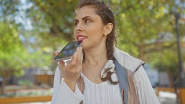 Beyaz kadın akıllı telefon sesli mesaj kullanıyor. Açık havada, yeşil bir parkta, gündüz vakti.