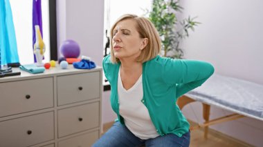 Rehabilitasyon kliniğinde arka planda fizyoterapi ekipmanıyla sırt ağrısı çeken olgun beyaz bir kadın..