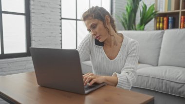 Dizüstü bilgisayarı üzerinde çalışan çok görevli genç bir kadın, ev içi aydınlık bir evde omuz ve kulak arasında duran bir telefonla çalışıyor..