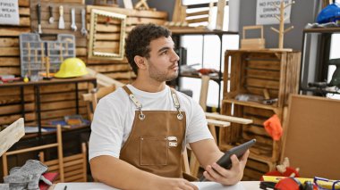 Marangozluk atölyesinde elinde tablet sakallı düşünceli bir adam. Etrafı aletlerle çevrili.