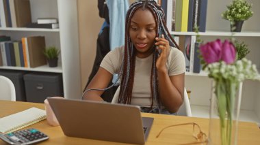 Örgü ören Afro-Amerikalı kadın ev ofisinin kurulumunda laptop ve telefon kullanıyor.