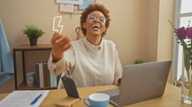 Süveter giymiş neşeli bir Afro-Amerikalı kadın elinde bir laptop ve masasında kahveyle evine uzanıyor..