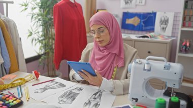A young muslim woman textile designer attentively using a tablet in a modern sewing atelier filled with fabric and sketches. clipart