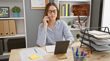 Gözlüklü genç esmer kadın organize iş yerinde telefon ve tablet kullanıyor.