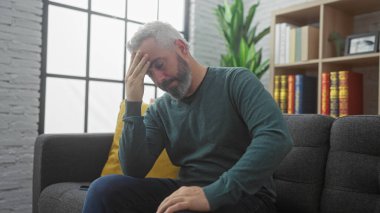 Stresli bir adam modern oturma odasında oturuyor. Alnı tutulmuş, endişe ve baş ağrısı belirtileri gösteriyor..