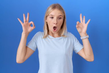 Günlük mavi tişört giyen genç beyaz kadın şaşırmış ve şok olmuş bir şekilde parmaklarıyla onay sembolü yapıyor. çılgın ifade 