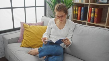 Modern oturma odasındaki rahat bir kanepede kitap okuyan genç bir kadın..