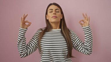 Genç İspanyol kadın çizgili tişörtle meditasyon yapıyor. Gözler kapalı, pembe arka planda.