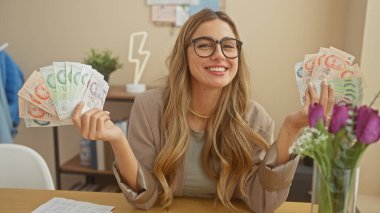 A smiling young woman in glasses holding singapore dollars indoors at home, portraying affluence and finance. clipart