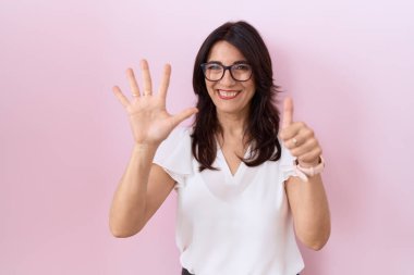 Orta yaşlı İspanyol kadın, günlük beyaz tişört ve gözlük takıyor. Altıncı parmaklarını gösterip işaret ederken kendinden emin ve mutlu bir şekilde gülümsüyor.. 