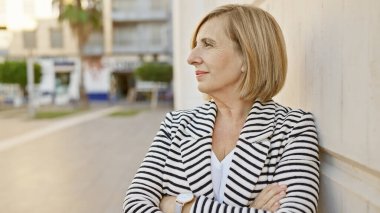 Çizgili ceketli, dengeli, orta yaşlı sarışın bir kadın güneşli bir şehir sokağında düşünceli bir şekilde duruyor..