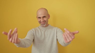 Süveterli sakallı yakışıklı kel bir adam, sarı bir duvara gülümsüyor, davetkar bir jest yapıyor..