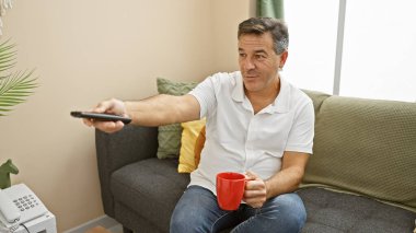 Orta yaşlı bir adam, rahat bir oturma odasında kahve içip uzaktan kumandayla rahatlıyor.