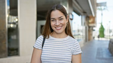 Mavi gözlü, esmer, gülümseyen bir kadın açık havada güneşli bir günün tadını çıkarıyor..