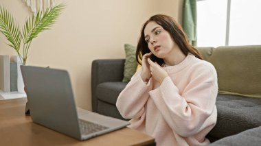 Pembe kazaklı düşünceli genç bir kadın. Akıllı telefon kullanıyor. Bilgisayarı açık ve arka planda kapalı bitkiler var..