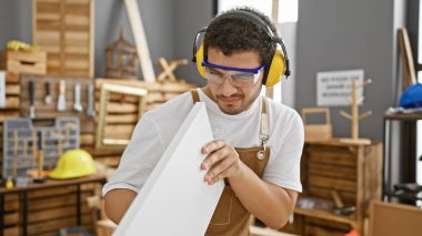 Güvenlik gözlükleri ve kulaklıkları olan genç adam iyi donanımlı bir marangozluk atölyesinde ahşabı inceliyor..
