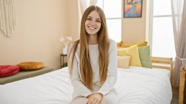 Gülümseyen genç bir kadın beyaz bir yatakta parlak bir yatak odasında renkli yastıklar ve modern bir dekorla oturuyor..