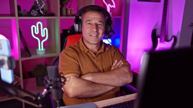 Middle-aged man with arms crossed smiles in a colorful, neon-lit gaming room at home. clipart