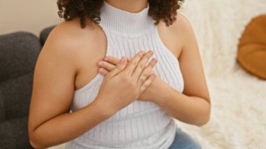 Kıvırcık saçlı genç bir İspanyol kadın ellerini sıcak bir oturma odasında göğsüne koyar..