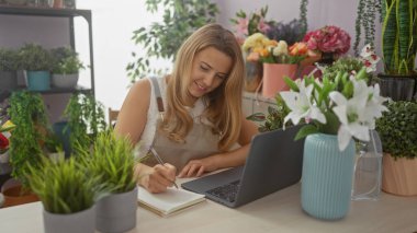 Gülümseyen genç bir kadın, çeşitli bitki ve buketlerle dolu canlı bir çiçekçide dizüstü bilgisayarına notlar alır..