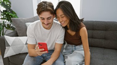 Irklar arası bir çift birlikte oturma odasında akıllı telefonlarıyla bir anı paylaşıyorlar, evde aşık bir erkek ve kadını resmediyorlar..