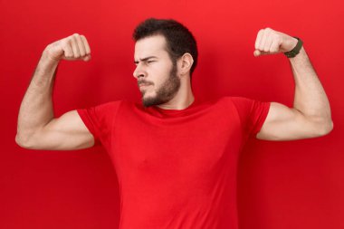 Sıradan kırmızı tişört giyen İspanyol genç adam gururla gülümseyen kol kaslarını gösteriyor. fitness konsepti. 
