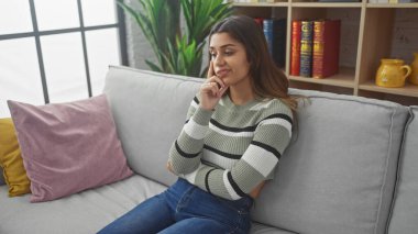 Düşünceli genç İspanyol kadın evdeki kanepede dinleniyor, gündelik tarzını ve modern iç güzelliğini sergiliyor..