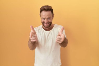 Sarı arka planda dikilen sakallı orta yaşlı adam mutlu ve komik yüzlü parmaklarını kameraya doğrultuyor. iyi enerji ve titreşimler. 