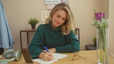 Genç sarışın bir kadın evindeki defterine çiçekler ve kulaklıklarla yazı yazıyor.