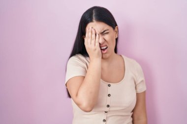 Young hispanic woman standing over pink background yawning tired covering half face, eye and mouth with hand. face hurts in pain. 