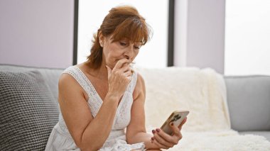 Düşünceli, orta yaşlı bir kadın bir koltuğa oturur, evde, akıllı bir telefon tutar, endişe veya tefekkür tasvir eder..