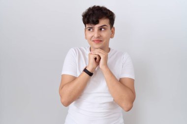 Gündelik beyaz tişört giyen, elleri çenesinde gergin ve heyecanlı bir şekilde yan tarafa bakan genç, çiftli olmayan bir adam. 