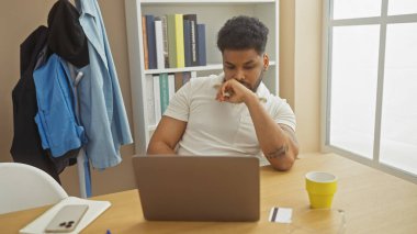 Evde dizüstü bilgisayarı, kahvesi ve akıllı telefonuyla çalışan düşünceli Afro-Amerikan bir adam.