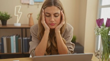 Düşünceli genç bir kadın, modern bir ev ofisinde dizüstü bilgisayarının başına oturmuş, stres ve tefekkür gösteriyor..
