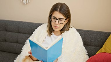 Odaklanmış genç bir kadın, rahat, modern bir oturma odasında kitap okur. Rahat ve çalışkan titreşimler yayar..