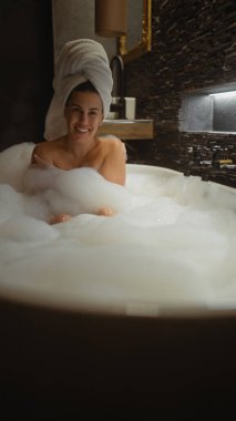 Kafasında havluyla gülümseyen genç bir kadın içeride lüks bir köpük banyosunda rahatlıyor..