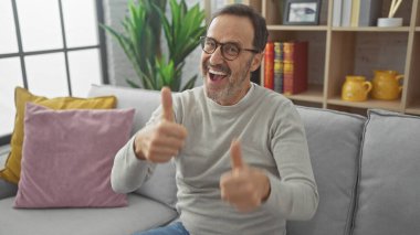 Gözlüklü, neşeli, orta yaşlı bir adam sıcak bir oturma odasında baş parmağını kaldırıyor..