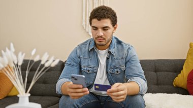 Genç bir adam, elinde akıllı telefon ve kredi kartı olan, muhtemelen internetten alışveriş yapan, modern bir oturma odasında bir koltukta oturuyor..