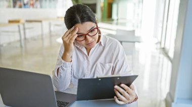 Latin kökenli genç bir kadın elinde tabletle bir ofis masasında otururken, arka planda dizüstü bilgisayar ve ışık dolu bir odayla endişeli görünüyor..