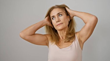 Üzerinde kolsuz bluz olan düşünceli sarışın bir kadın beyaz bir arka plan karşısında elleri başının arkasında duruyor..