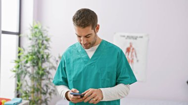 Yeşil önlüklü profesyonel bir İspanyol, arka planda anatomik çizelgeler olan bir klinik odasında akıllı telefon kullanıyor..