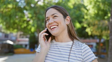 Güneşli yeşil parkta akıllı telefondan konuşan gülümseyen genç kadın.