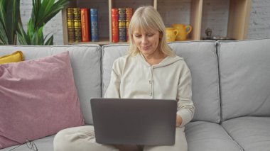 Sarışın bir kadın laptopta çalışıyor. Kapalı bir koltukta oturuyor. Rahat ve odaklanmış bir ortam yayıyor..