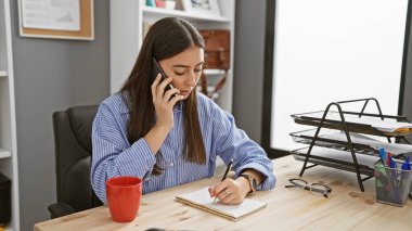 Genç bir İspanyol kadın, modern bir ofis ortamında not alırken bir telefon görüşmesi yapıyor..