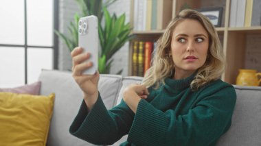 Kafkasyalı genç bir kadın akıllı telefonuyla evde selfie çekiyor. Oturma odasını sergiliyor..