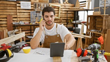 Sakallı genç bir adam ahşap atölyesinde telefonla konuşuyor. Etrafı alet ve kerestelerle çevrili..